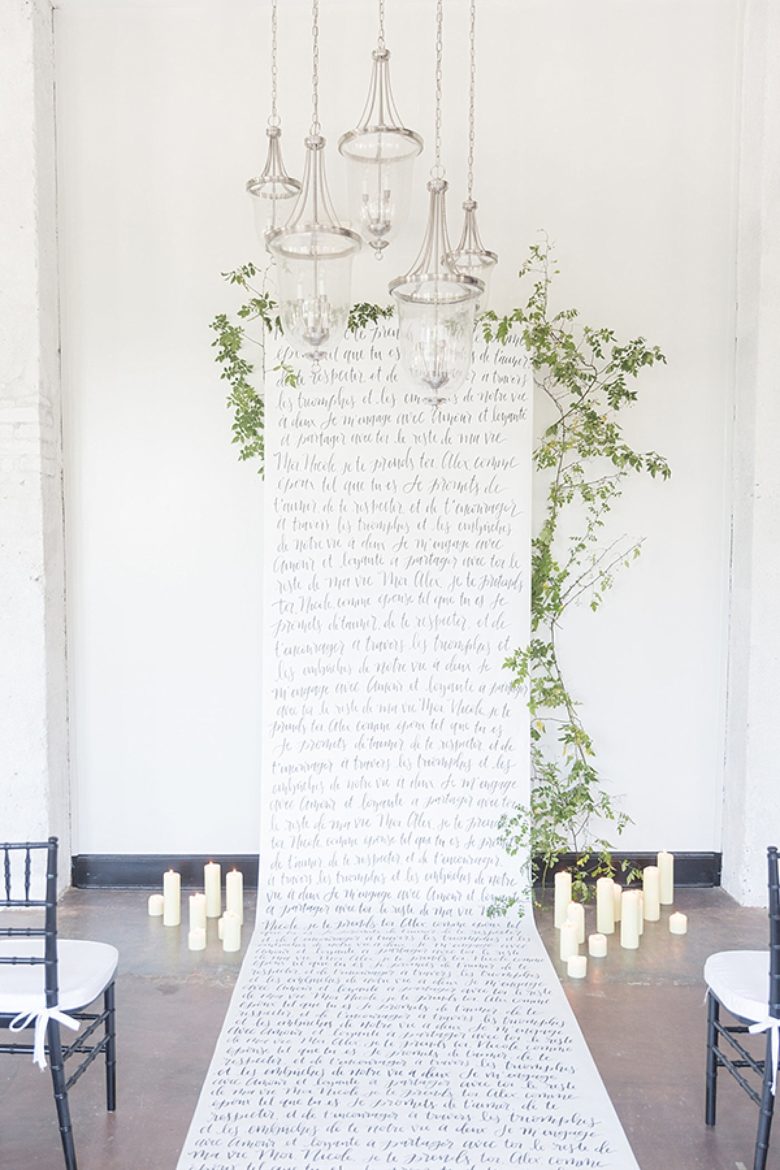 long handwritten-style scroll as backdrop to altar as well as aisle, bordered by white pillar candles on the ground and clear glass pendant lights handing above