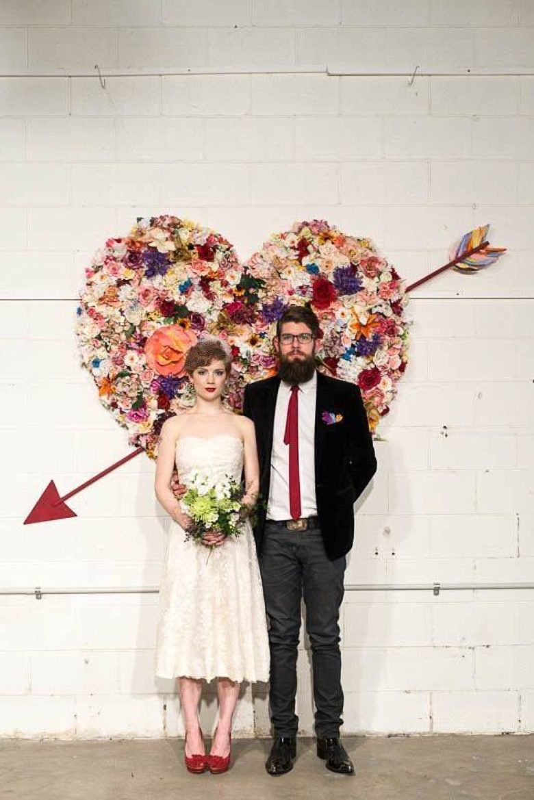 bride and groom stand in front of heart made of a flower collage, with an arrow through it