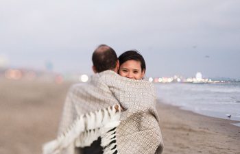 A couple embrace on a beach, wrapped up in a blanket