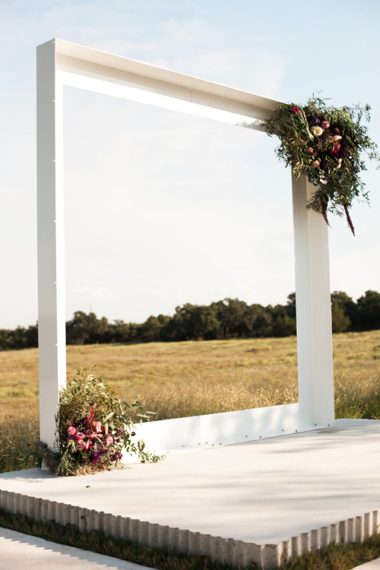 large white square frame on a short raised cement altar, adorned with flowers in two opposite corners, in a field