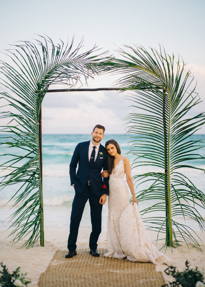 bride and groom at altar on beach framed by tall leafy fronds