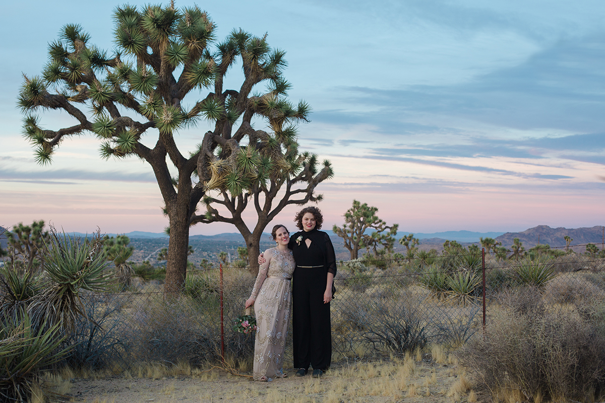 A women holding a bouquet in a long dress embroidered with silver designs and taller woman in long black dress have their arms around each other and are standing in the desert in front of a Joshua Tree in a Gina Clyne photo