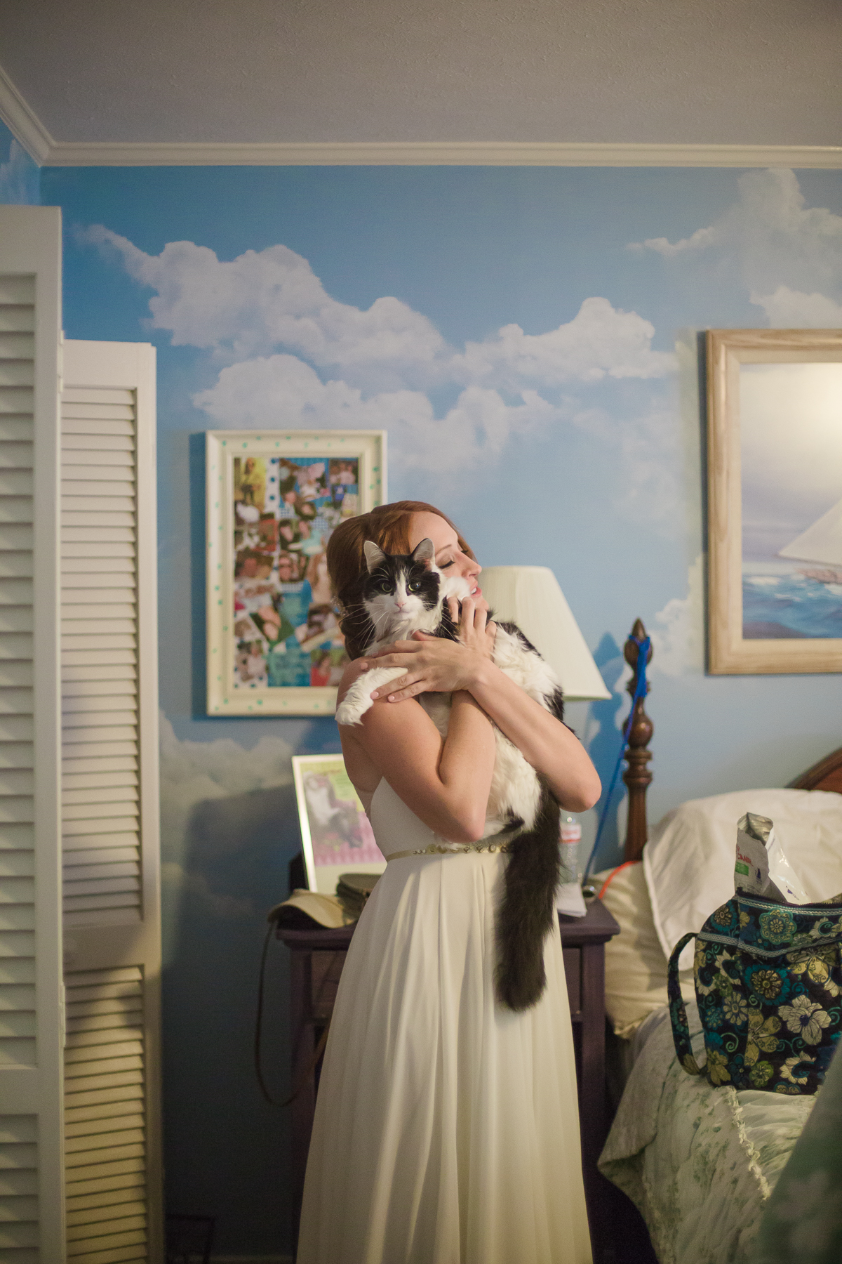 bride holds grey and white cat on her shoulder while standing in bedroom with walls painted with clouds in a Gina Clyne photo