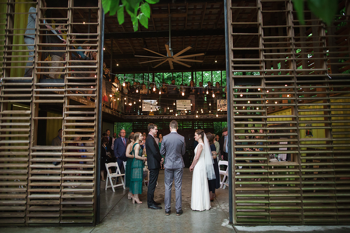bride and groom and officiant stand with their backs to camera during a wedding ceremony taking place in two-story barn with dramatic louvered wooden walls in a Gina Clyne photo