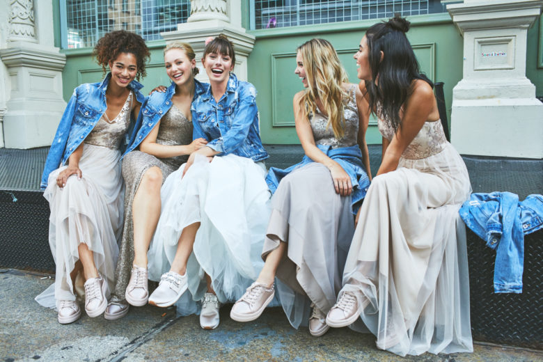 Group of bridesmaids and bride sitting together laughing wearing denim jackets over their gowns with matching sneakers.