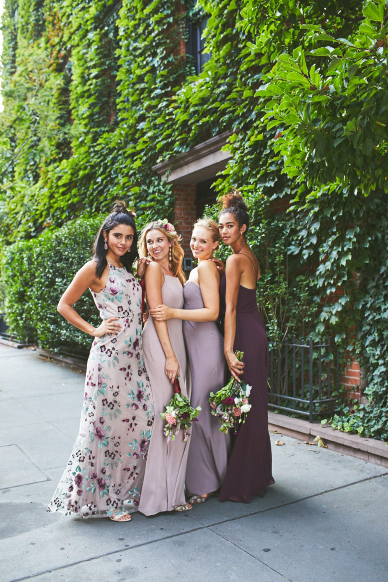 Group of bridesmaids wearing mismatched dresses in a floral and purple color palette against an ivy covered wall.