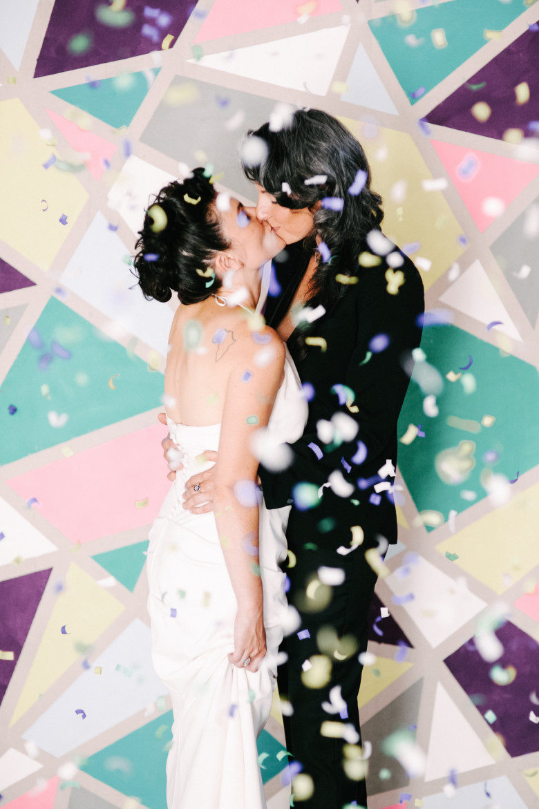 two women in wedding finery embrace amid confetti in front of geometric painted backdrop
