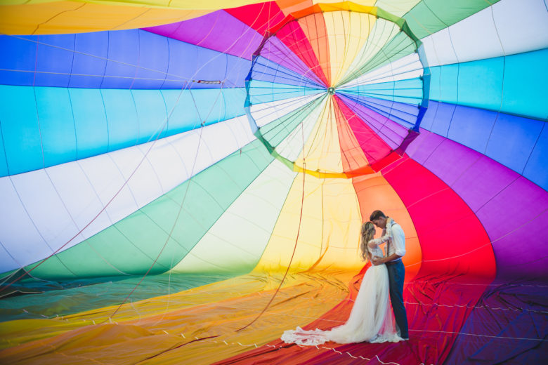 couple in wedding clothes stand in embrace insight a rainbow hot air balloon