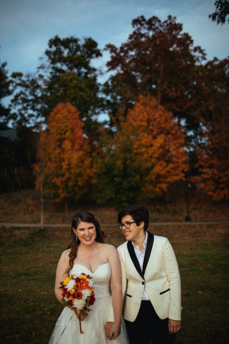 Wedding couple holding hands, smiling, in front of trees with autumn foliage