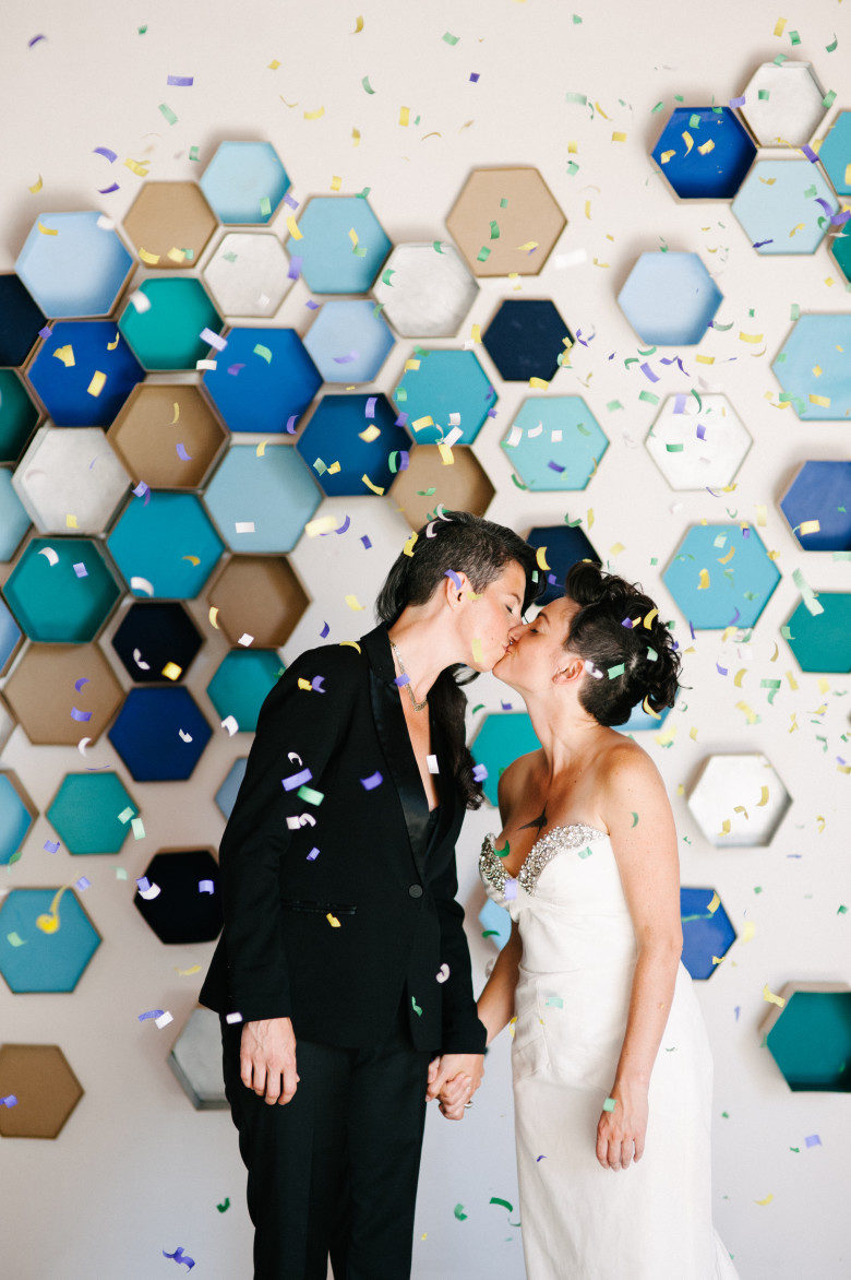 two women in wedding finery kiss amid confetti in front of dimensional honeycombed style geometric backdrop
