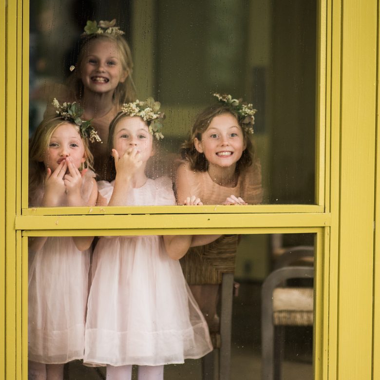 four young girls in pink dresses and flower crowns smile out through a rain-drenched yellow framed window