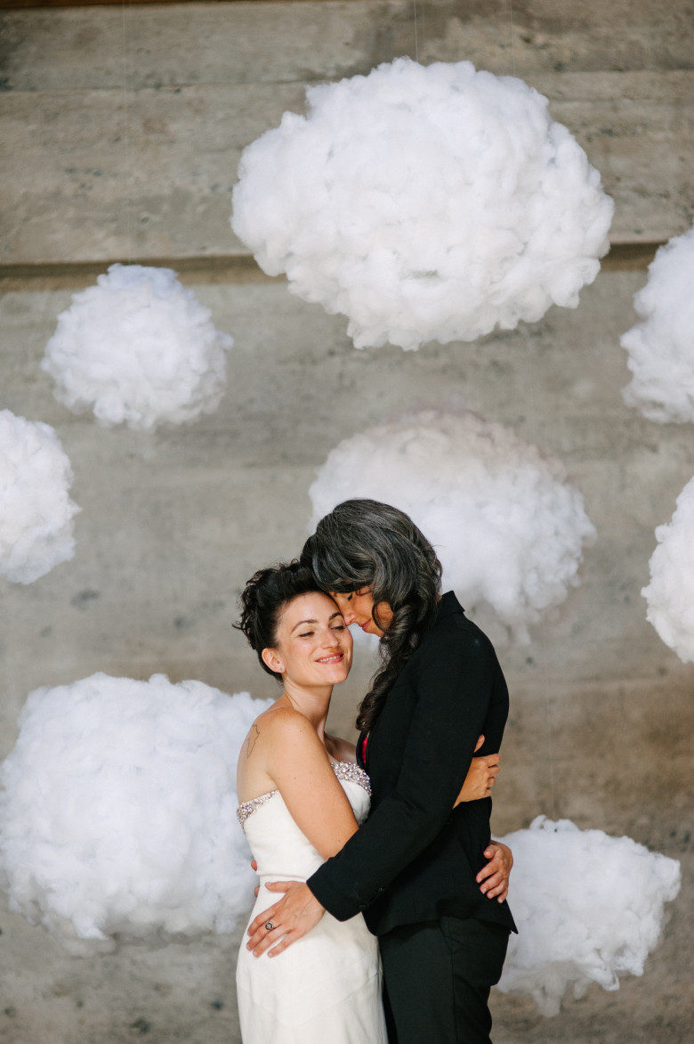 two women in wedding finery embrace amid fluffy white clouds in front of industrial style cement wall