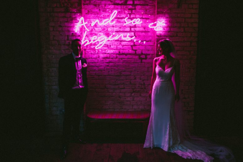bride and groom in front of pink neon-style sign that reads, "And so it begins..."