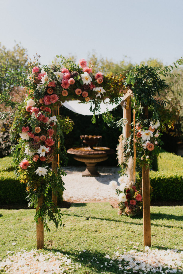 chuppah trimmed with lush greenery and pink and white flowers in a garden in front of a fountain
