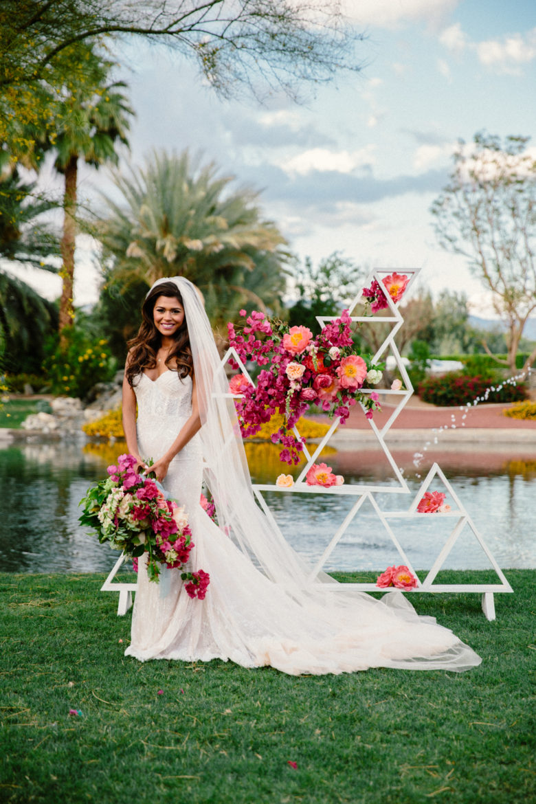 smiling bride holding large bouquet of cascading bougainvillea stands in front of a white triangle collage adorned with pink flowers, in front of a lake or pool in a tropical setting