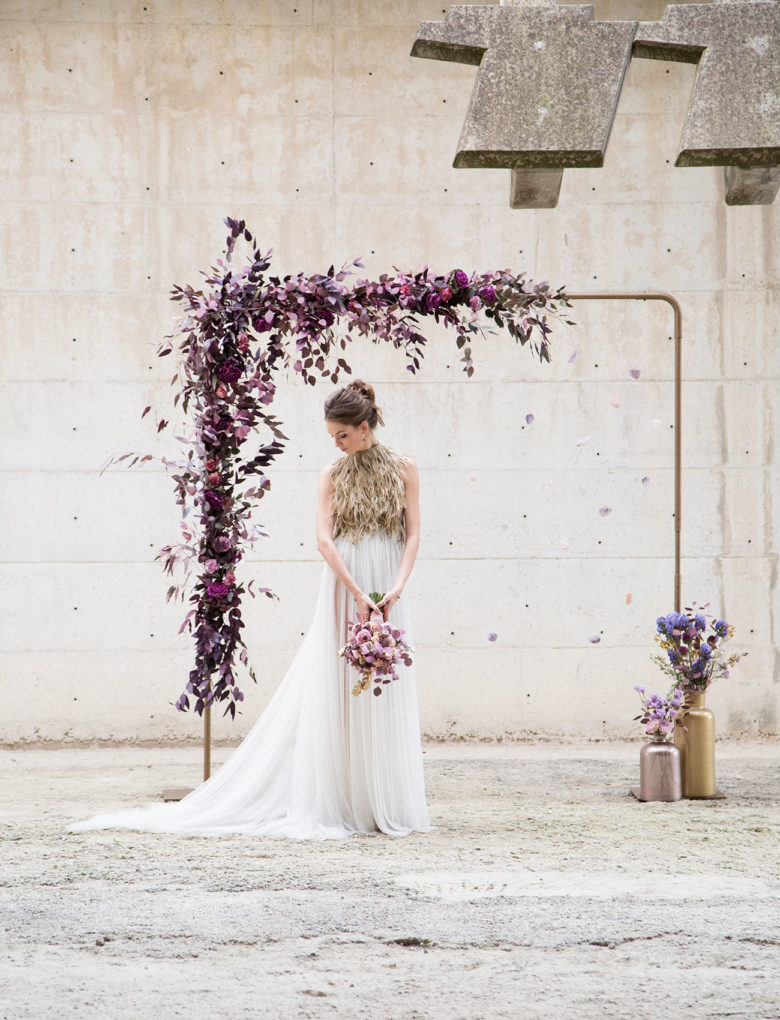 bride stands in front of a square frame lined on one corner with purple flowers, with two vases of purple flowers in the opposite corner, in front of a cement wall