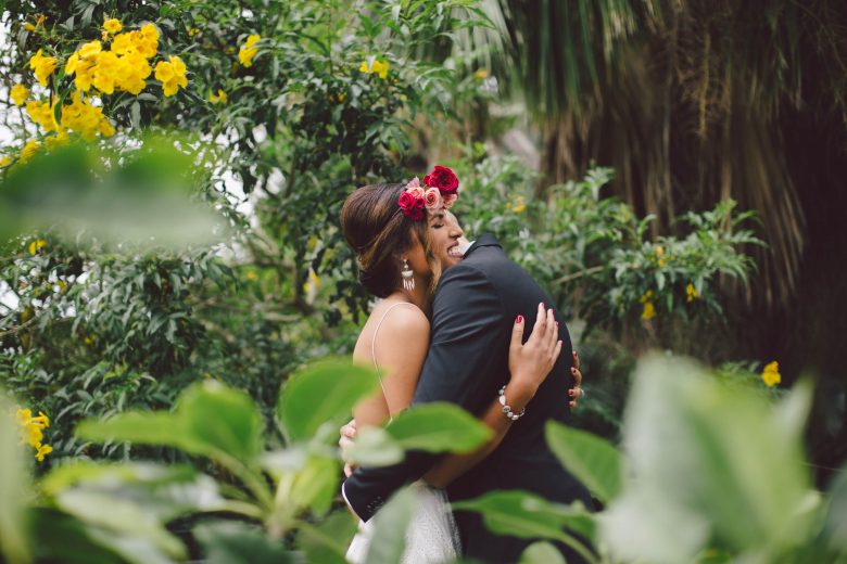 Bride with red and pink flower crown smiles and embraces groom in a lush tropical environ