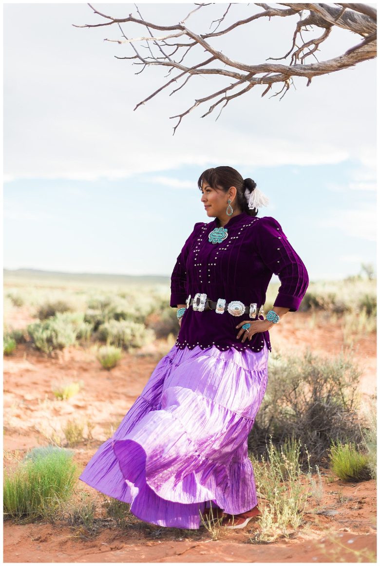 Woman in purple top and long skirt with silver belt and silver and turquoise accessories in a desert under an empty tree branch