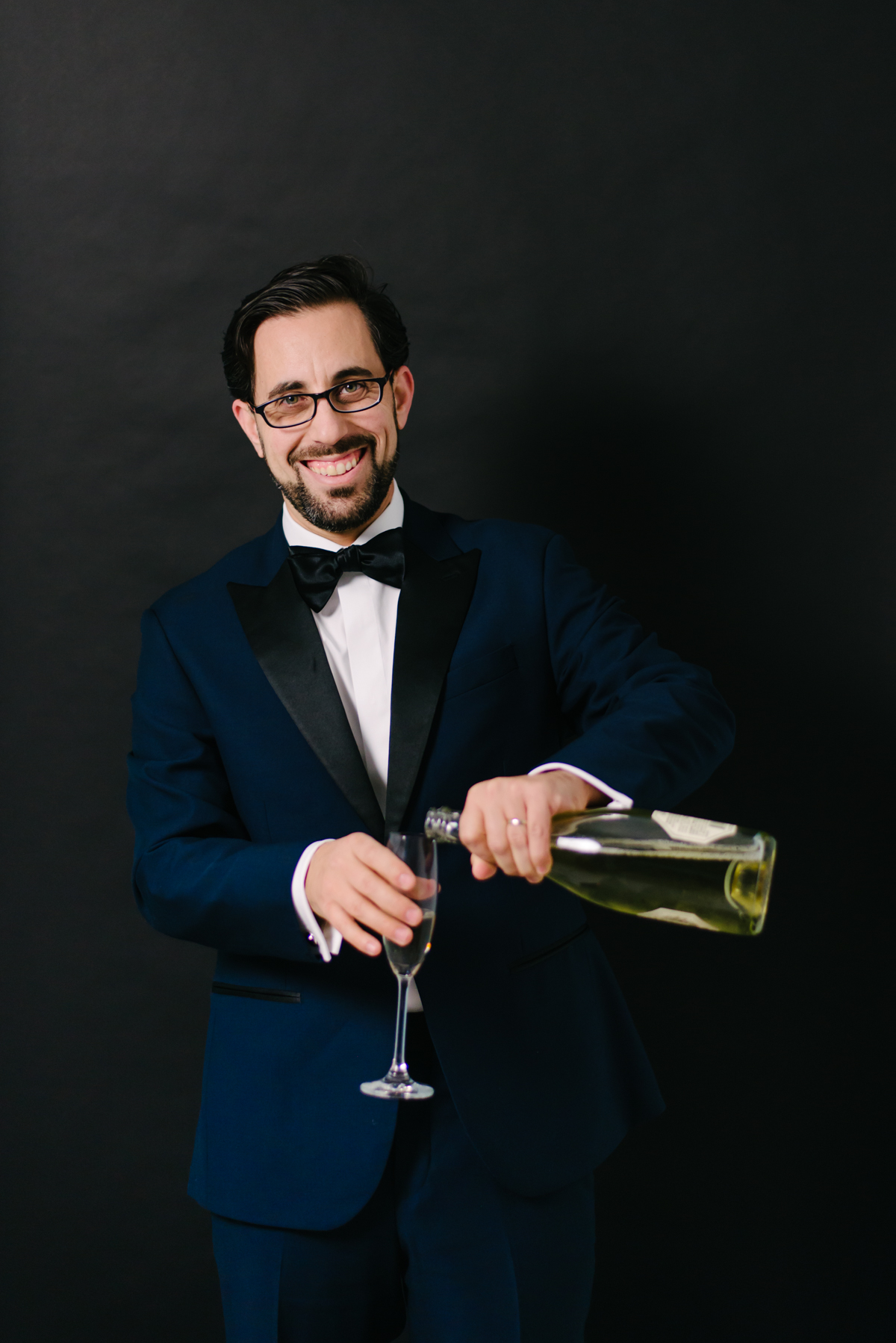 A ridiculously good looking man wears a dark blue tuxedo while pouring wine into a glass