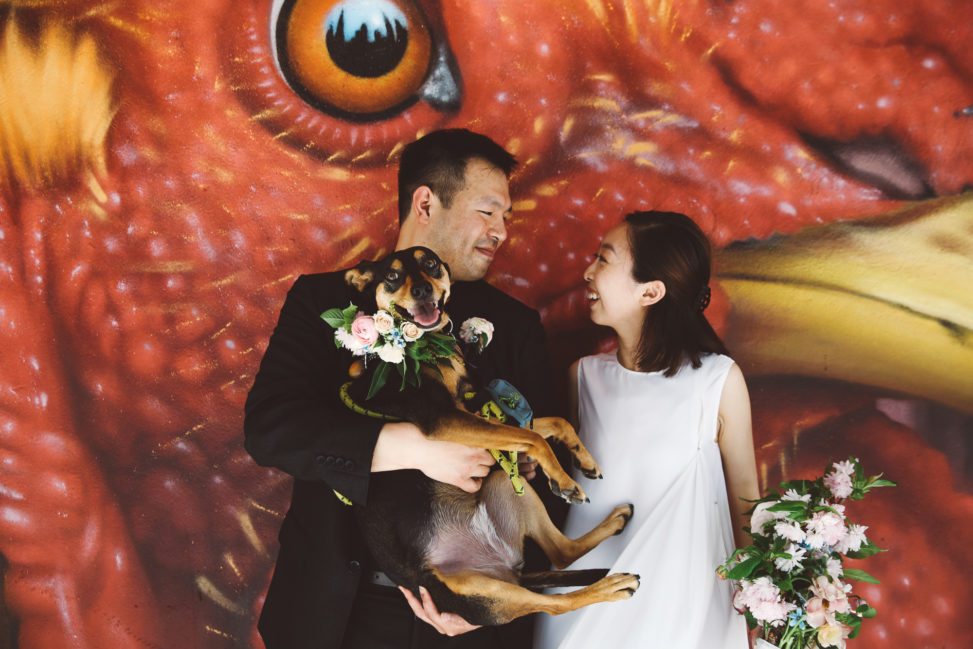 Couple smiles at each other in front of a giant mural of a red bird with yellow beak, the skyline reflected in its pupil. The groom holds a dog with a flower wreath collar; the bride holds a corresponding bouquet.
