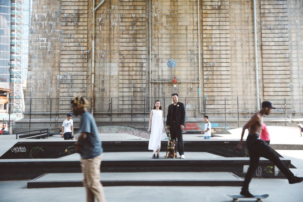 Couple with dog on leash stands in the middle of a skate park as skateboarders whiz by