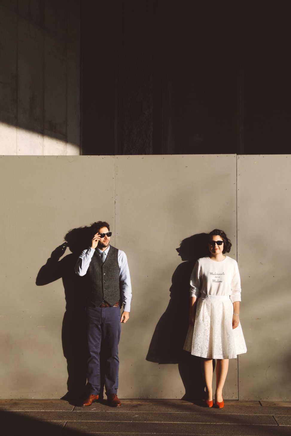 Couple in sunglasses stand in front of a temporary wall outside, the sun creating dramatic elongated shadows on the wall behind them