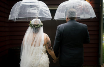 Couple in wedding attire holding hands with their backs to the camera, standing under clear plastic umbrellas