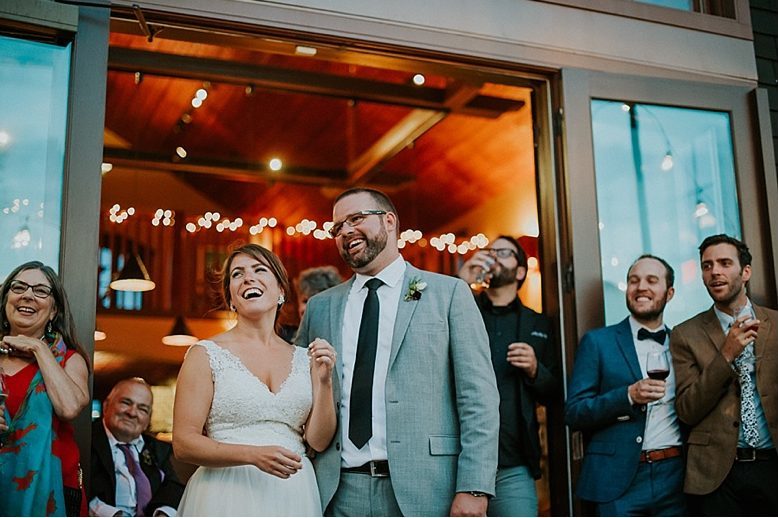 laughing bride and groom standing in front of open door to warmly lit reception