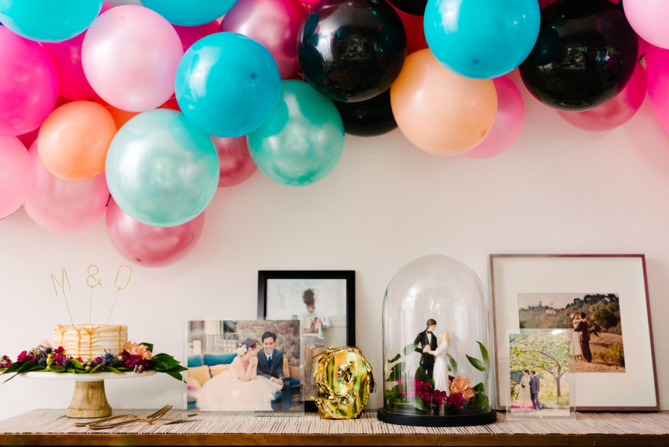 vintage credenza with wedding photos in clear acrylic block frames from crate and barrel, a vintage cake topper in a cloche, and caramel drip wedding cake on a crate and barrel cake stand, with balloons floating overhead