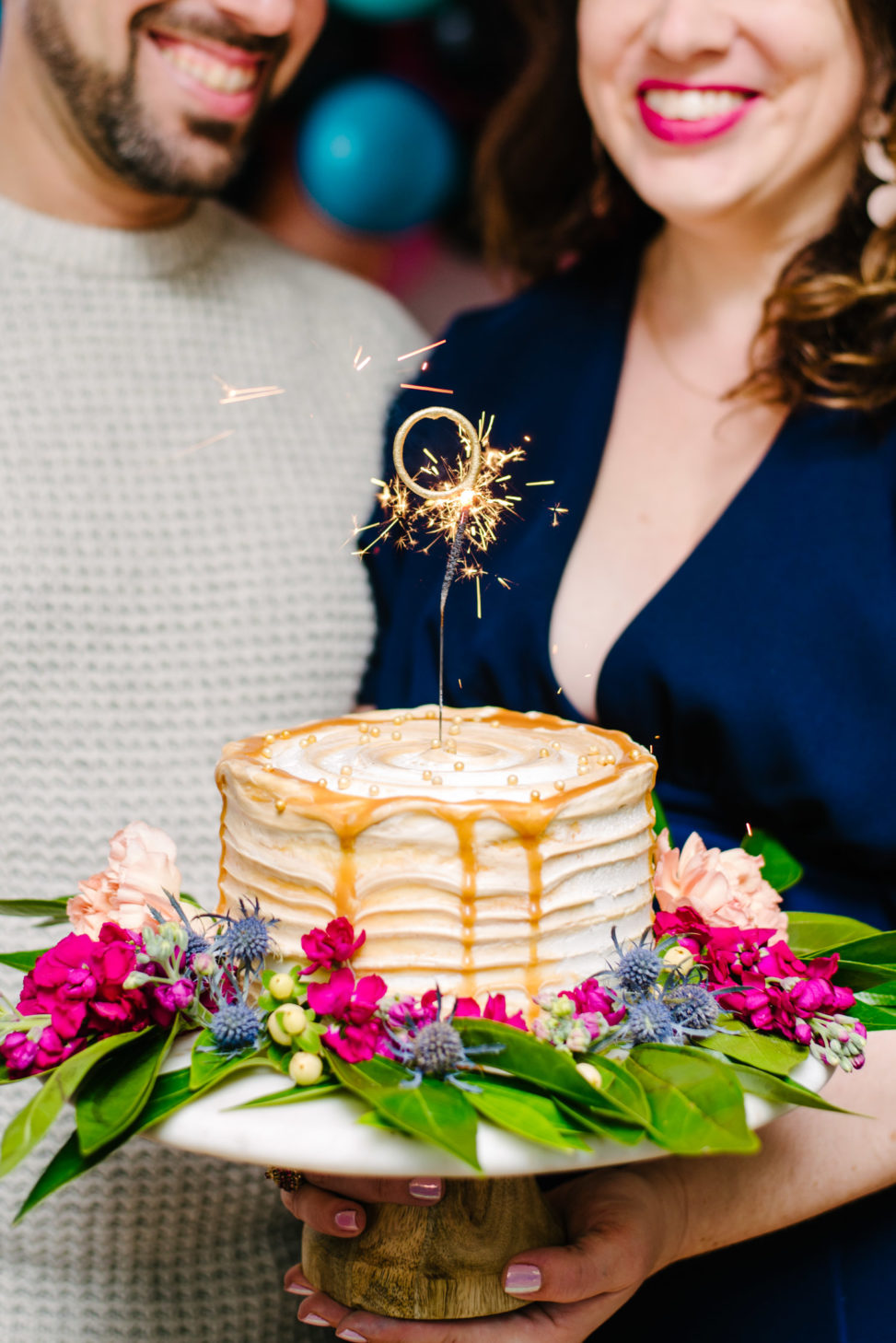 couple holding caramel drip cake on a crate and barrel cake stand with sparkler candle that says the number nine