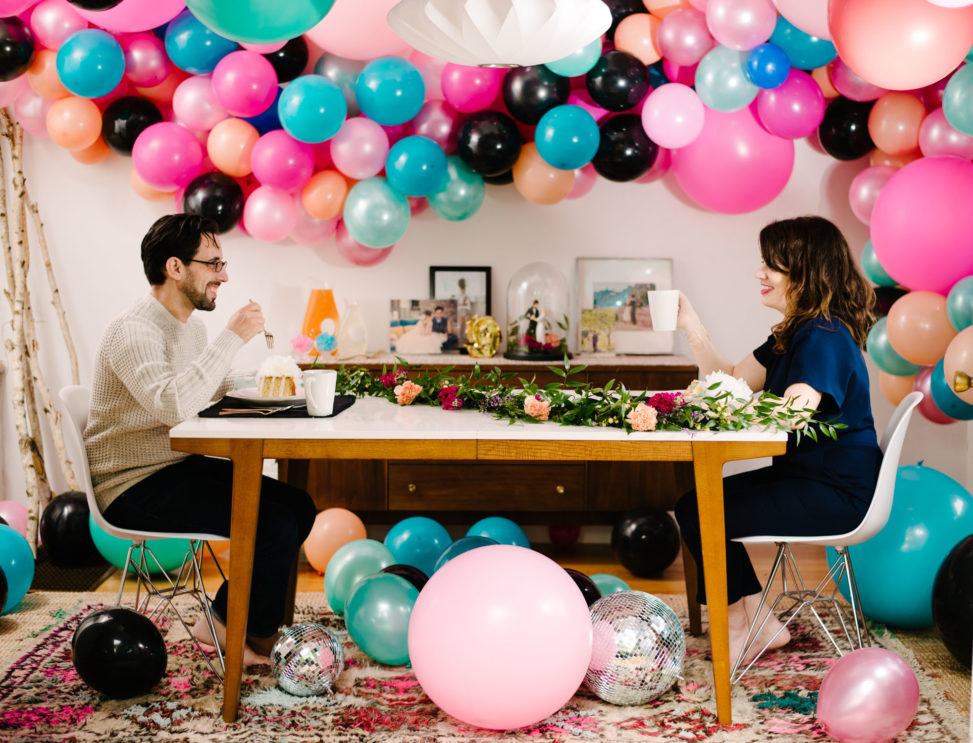 couple sitting at a breakfast table eating wedding cake in a room filled with disco balls and brightly colored balloon installation