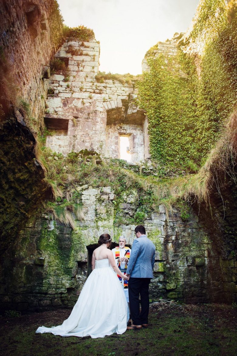 bride and groom holding hands in front of officiant in a chamber of castle ruins