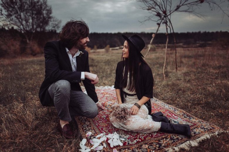 bride with black fedora and groom in jeans sitting on a picnic blanket in a field