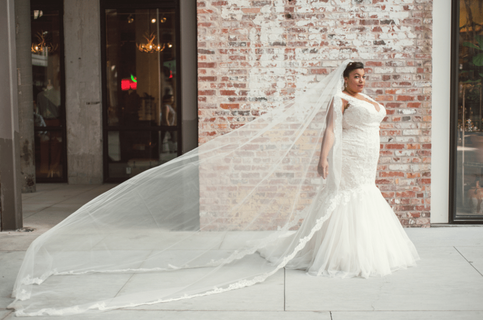 plus size black bride in front of a brick wall in a mermaid wedding gown and cathedral length veil blowing behind her in a photo by You Are Raven
