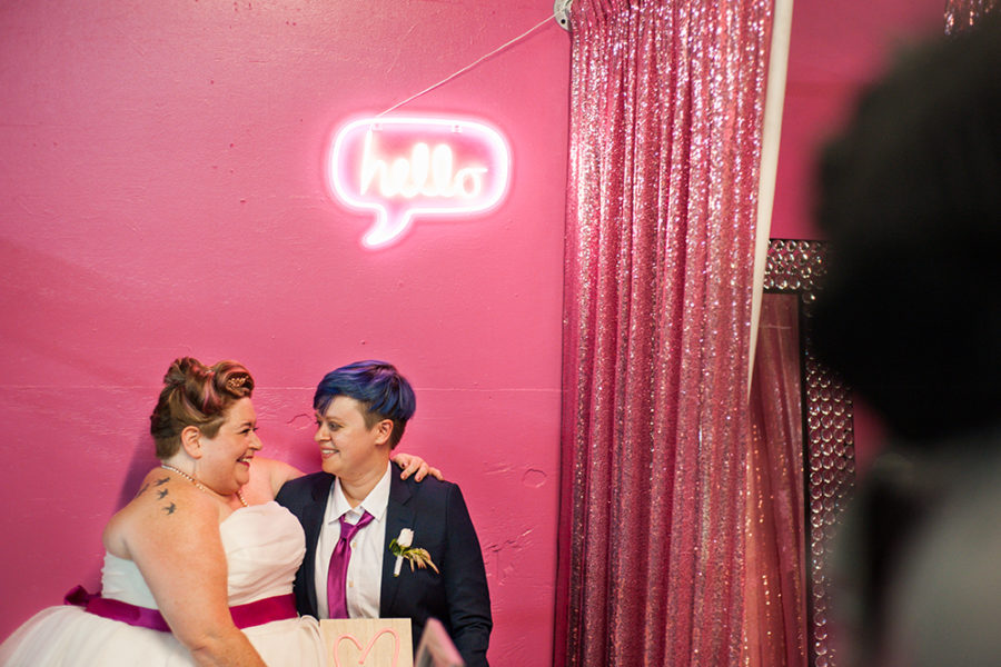 A queer couple in a suit and wedding dress sit in front of a pink wall beneath a neon sign that reads "Hello" in a photo by You Are Raven