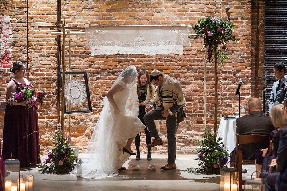 Bride and groom stomp on glasses at altar in front of an exposed brick wall in a photo by You Are Raven