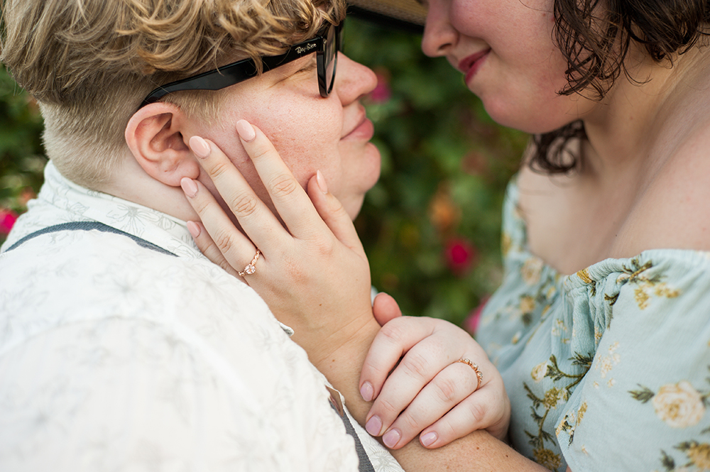 close-up of couple gazing into each other's eyes as one, who's wearing a floral dress, is touching the other, who is wearing black glasses, on the cheek in a photo by You Are Raven