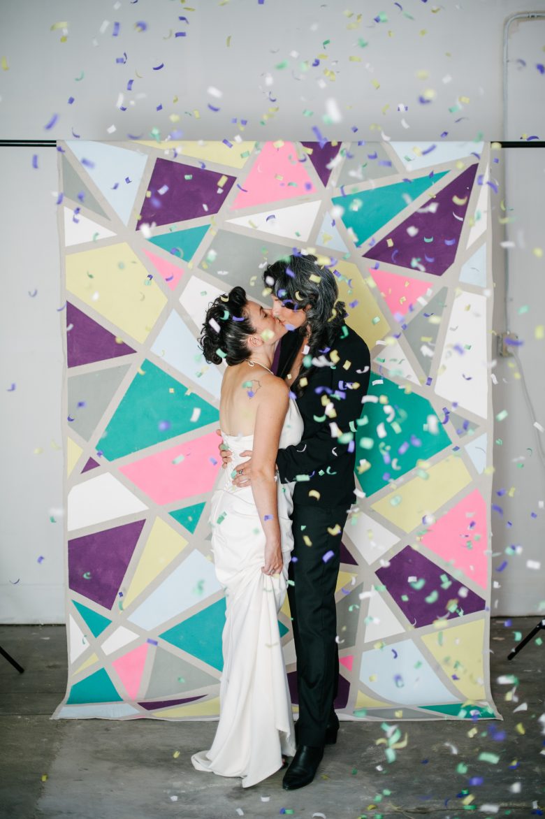 lesbian couple standing in front of a diy painted backdrop with confetti falling