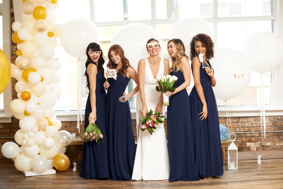 bride in white holding pink and green bouquet surrounded by bridesmaids in floor-length navy dresses, some wear silly glasses, others hold props for the photo