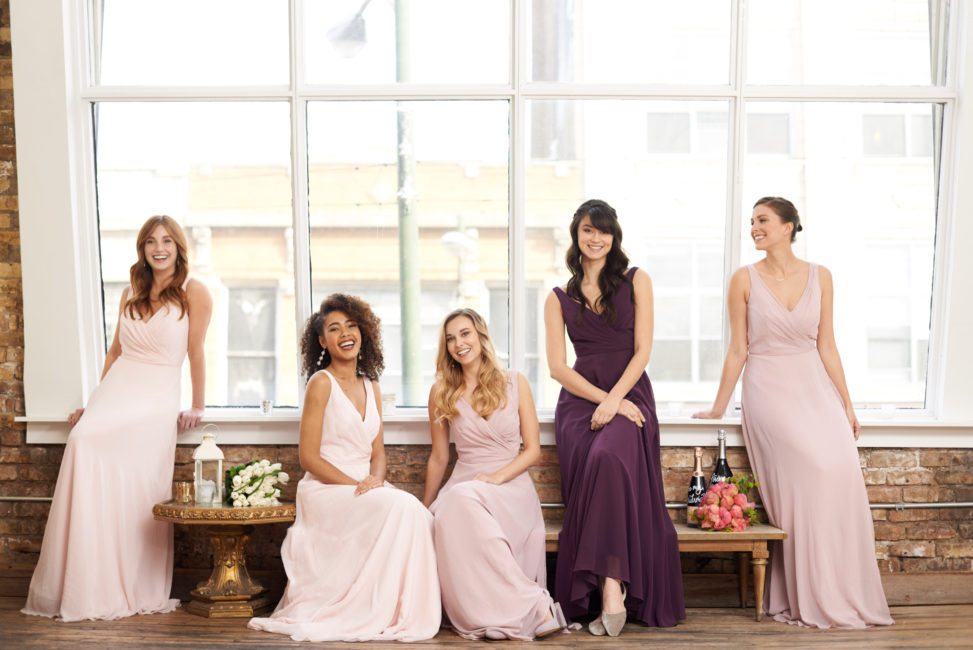 bridesmaids in pink or purple floor-length dresses, sitting in front of a brightly lit loft window