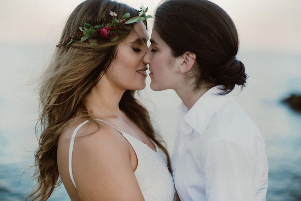 lesbian couple, one with floral crown, kissing on a beach