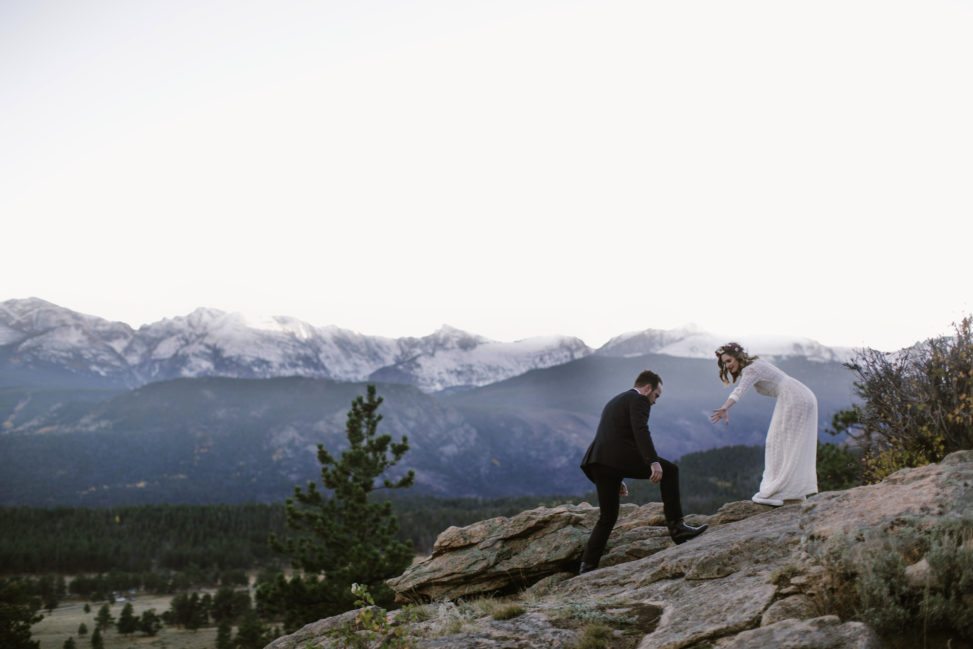 bride and groom climbing rocks in front of white mountain peaks