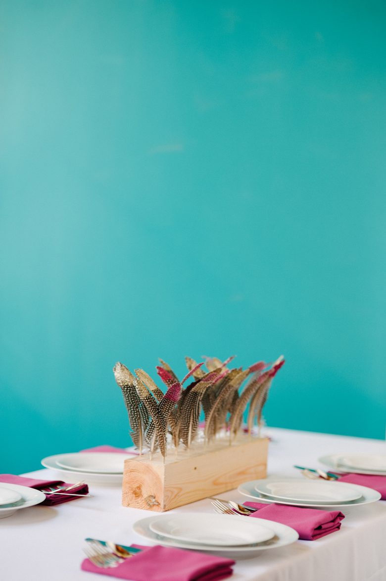diy wood block wedding centerpiece with painted gold feathers and hot pink napkins in front of a teal wall