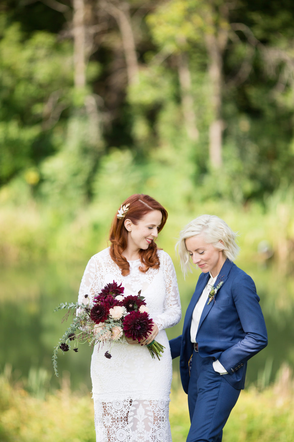 bride in lace gown and bride in blue suit standing in front of trees