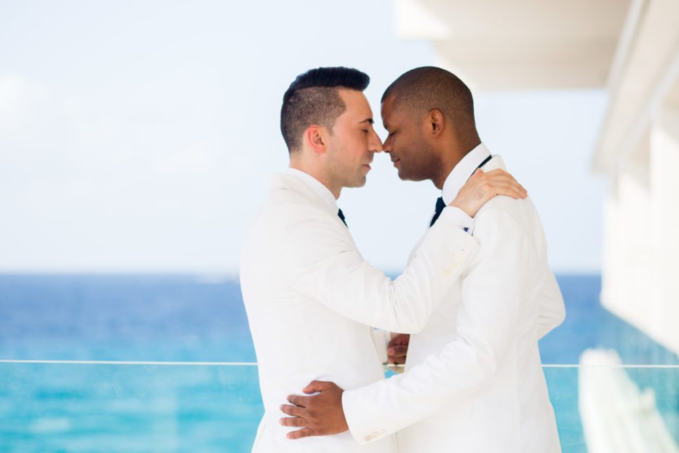 two grooms in white suits embracing in front of blue ocean