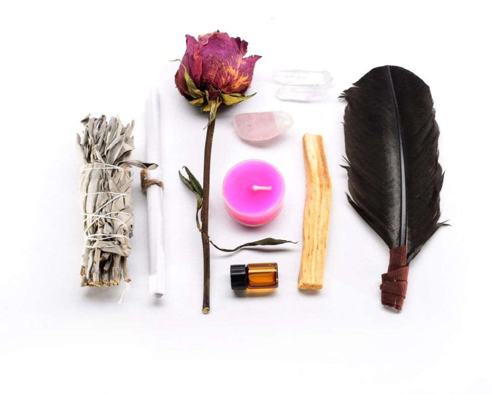 Rose, candle, feather, bottle, crystals, paper roll, wood piece, and bundle of dried leaves on white background