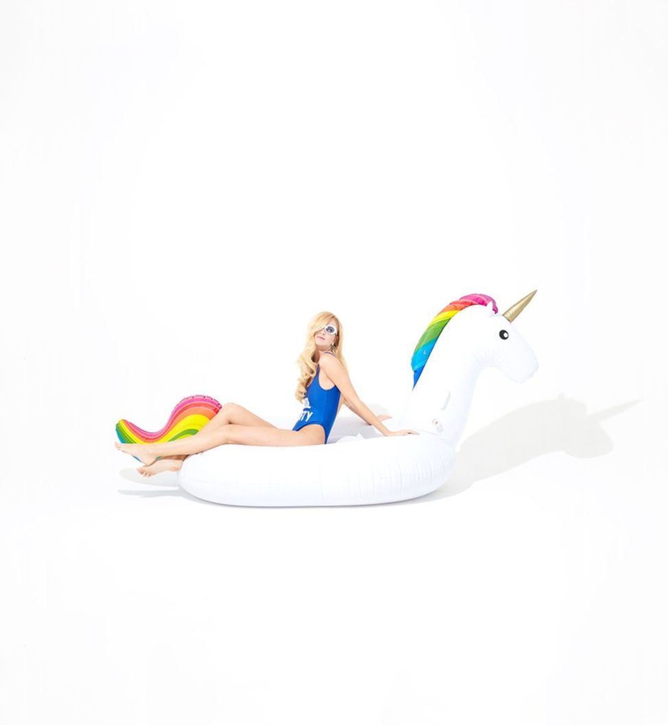 Blonde woman in swimsuit sitting in pool floaty of unicorn with rainbow hair