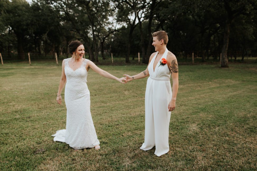 lesbian couple holding hands in a field, wearing a wedding dress and a white BHLDN jumpsuit