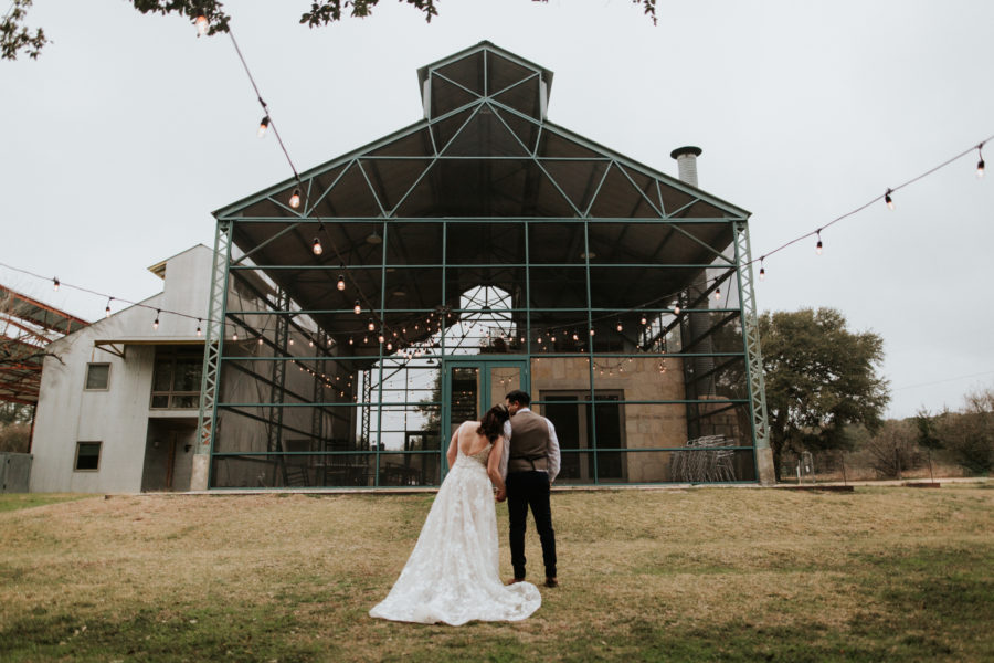 couple in wedding outfits standing in front of an indoor-outdoor event space, leaning on one another under string lights