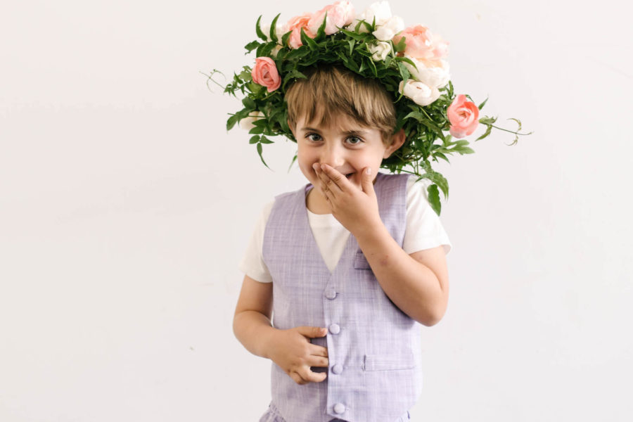 Young boy in a lavender vest and short-sleeved shirt wears a large flower crown and giggles behind his hand